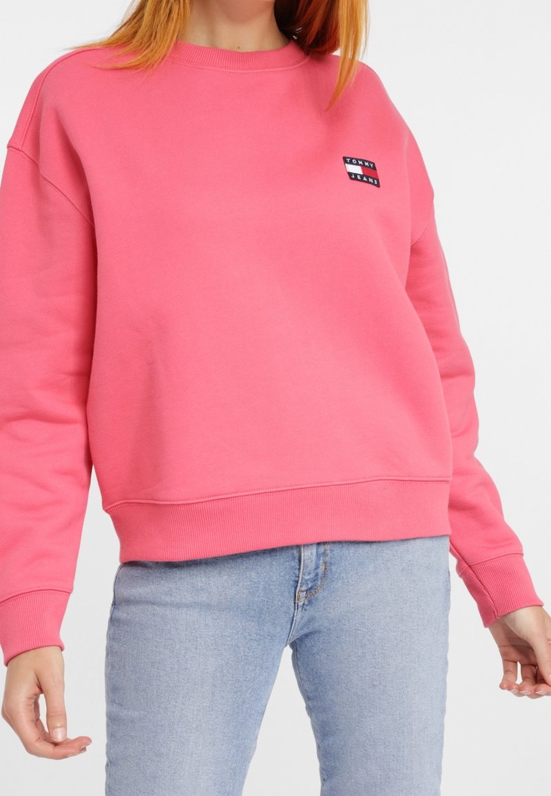 Femme portant un sweat-shirt Tommy Jeans rose vif avec des poignets et une ceinture côtelés, associé à un jean en denim bleu clair.