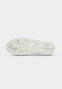 Semelle de chaussure en caoutchouc blanc avec des motifs de bande de roulement diagonaux et en chevrons, avec le nom de la marque "GUESS" embossé au centre.
