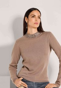 Vrouw met lang donker haar, gekleed in een lichtbruin trui met metalen details op de kraag, poseert met één hand op haar heup en de andere in de zak van haar spijkerbroek.