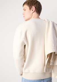 Junger Mann in cremefarbenem geripptem Pullover, der eine beige Jacke über der Schulter hält, trägt helle blaue Jeans und sieht nach links vor einem einfarbigen Hintergrund.