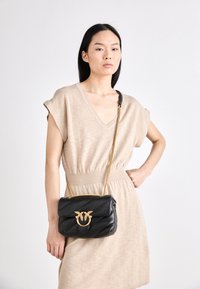 Sac en cuir matelassé noir avec quincaillerie dorée et une bandoulière en chaîne, présentant un logo unique entrelacé à l'avant.