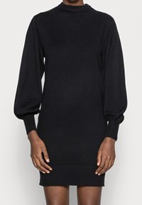 Robe noire en maille avec un col montant, des manches longues bouffantes et des poignets côtelés. L'ourlet est au-dessus du genou et présente une texture lisse.