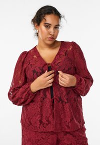 Bordeaux blondes bluse med udblæste ærmer, blomstermønster, dyb V-hals og en snøre lukning foran. Teksturen ser delikat og gennemsigtig ud.