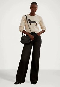 Γυναίκα με κρεμ πουλόβερ με γραφικό σχέδιο ντάκσχοντ, μαύρες φαρδιές παντελόνες, μαύρη τσάντα ώμου και μαύρα παπούτσια, που στέκεται μπροστά σε λευκό φόντο.