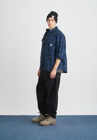 Μοντέλο που φοράει ένα καρώ φανελένιο πουκάμισο σε μπλε και μαύρο χρώμα, μαύρα παντελόνια, μπεζ μπότες και ένα μαύρο σκούφο. Χαρακτηριστικά περιλαμβάνουν μια τσέπη στο στήθος και μανίκια που είναι τυλιγμένα.