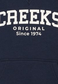 Marineblaues Sweatshirt mit "CREEKS ORIGINAL Seit 1974" in großen weißen Buchstaben. Verfügt über einen V-Ausschnitt und eine Tasche auf der Vorderseite.