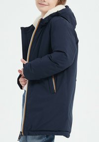 Veste matelassée bleu marine avec une capuche doublée de fourrure, dotée d'une fermeture éclair orange, de poches latérales et d'une coupe décontractée. Texture douce et design chaud.