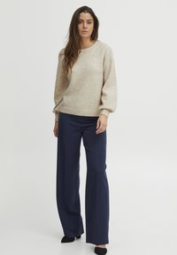 Beige neulepusero, jossa on tekstuuri ja koristelut, yhdistettynä tummansinisiin leveisiin housuihin. Mustat kengät viimeistelevät asun.