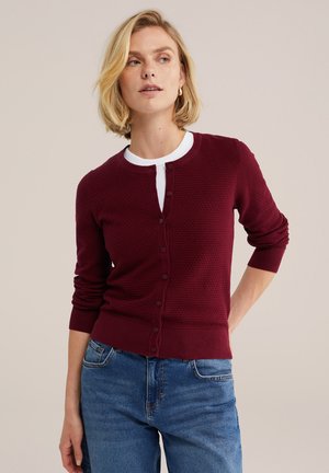 Žena s krátkymi blond vlasmi, ktorá nosí zapínaný vínovo textúrovaný sveter cez biely top a modré džínsy, stojí s jednou rukou za chrbátom.