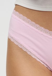 Κοντινό πλάνο ενός ατόμου που φοράει ανοιχτό ροζ βαμβακερά εσώρουχα με δαντέλα κατά μήκος της μέσης και των ανοιγμάτων στα πόδια.