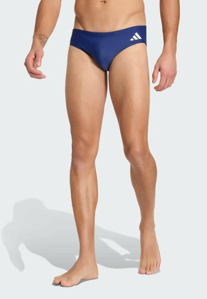 Male swimmer wearing navy blue briefs with white Adidas logo, standing barefoot against a plain light background.