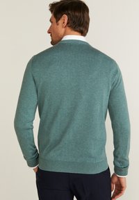 Tealfärgad stickad tröja med rund halsringning, ribbade ärmslut och nederkant, med en slät textur och en klassisk passform, som bärs över en skjorta med krage.