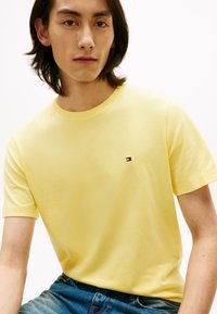 Yellow cotton T-shirt with short sleeves, crew neckline, and a small embroidered logo in red, white, and blue on the chest. Blue jeans visible.