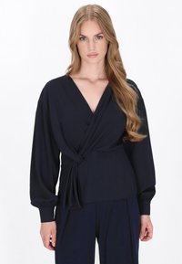 Top avvolgente navy con maniche lunghe, realizzato in tessuto morbido. Presenta un profondo scollo a V e un dettaglio attorcigliato in vita. Texture morbida e fluida.