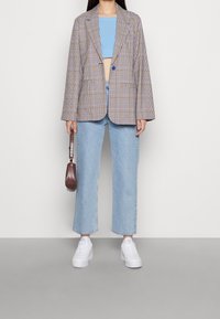 Osoba s kostkovaným blejzrem přes modrý crop top, světle modrými širokými džíny, bílými teniskami a malou hnědou kabelkou držící ji u boku.