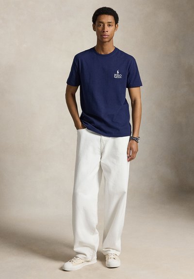 Νέος άντρας στέκεται με το ένα χέρι στην τσέπη, φορώντας μπλε σκούρο Polo Ralph Lauren μπλουζάκι, λευκό φαρδύ παντελόνι, λευκά sneakers και ρολόι χειρός.