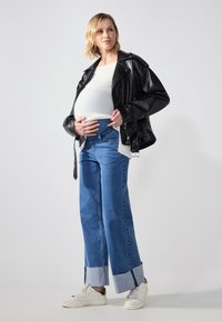 Jaqueta de pele negra sobre uma blusa branca, combinada com jeans de perna larga azul com detalhes em denim e azul claro na barra. Ténis brancos são usados.