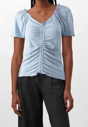 Kobieta ubrana w jasnoniebieski prążkowany top z marszczonym środkiem i krótkimi bufkami, w połączeniu z ciemnymi plisowanymi spodniami.