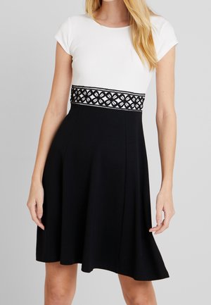 Kobieta w sukience do kolan z białym dopasowanym górnym dołem, czarną spódnicą oraz czarnym koronkowym paskiem z kwiatowym wzorem w talii.