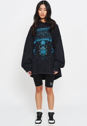 Oversized zwarte hoodie met blauwe grafische print van een kever en hemelse motieven. Gekleed met zwarte fietsshorts en hoge sneakers.