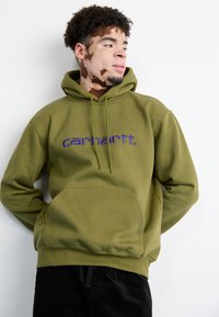 Felpa verde oliva in tessuto morbido, con tasca frontale e logo "carhartt." in viola. Cappuccio con coulisse regolabile.