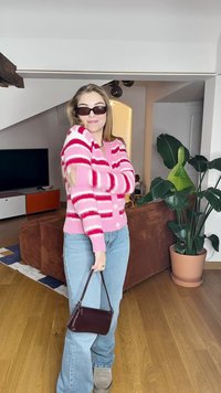 Cardigan às riscas rosa e branco com botões, usado sobre jeans azul claro. O acessório inclui uma pequena bolsa castanha. Piso de madeira e plantas de interior.