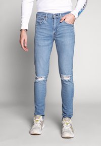 Světle modré slim džíny s roztržením na kolenou, vyrobené z denimu, v kombinaci s bílými a šedými teniskami. Mají středně vysoký pas a kapsy.