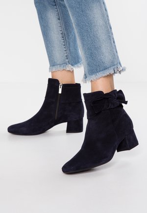 Bottes en daim bleu marine longueur cheville avec talons blocs, fermeture éclair latérale et détail nœud, portées avec un jean bleu clair à ourlet effiloché.