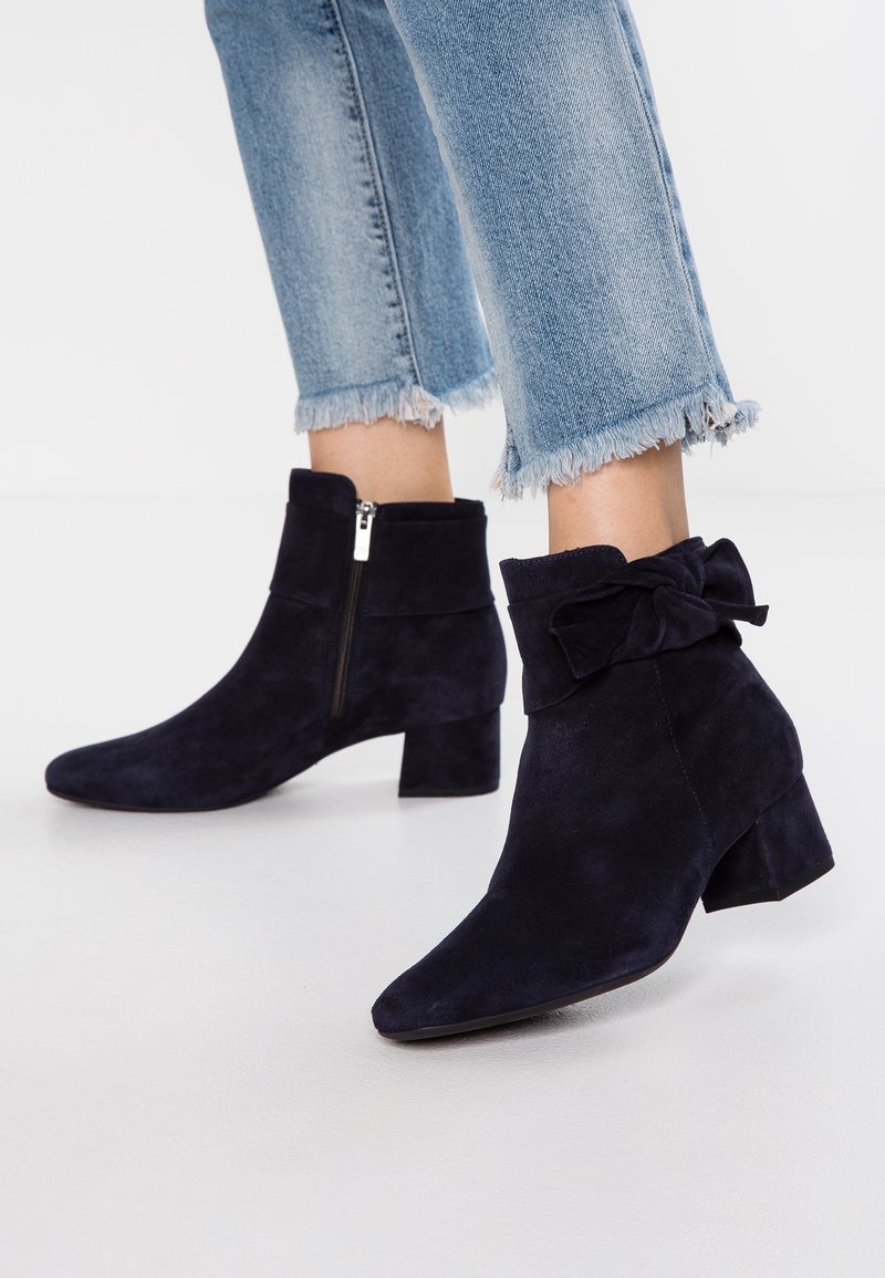 Bottes en daim bleu marine longueur cheville avec talons blocs, fermeture éclair latérale et détail nœud, portées avec un jean bleu clair à ourlet effiloché.