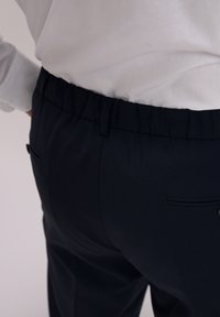 Námořnické kalhoty s elastickým pasem a jednou zadní kapsou. Tkanina je hladká s mírným leskem. Bílá košile částečně viditelná nahoře.