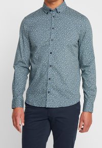 Camisa de manga larga de tela en verde azulado oscuro con un pequeño patrón floral en blanco y azul claro, con cuello de botonadura y botones en la parte delantera.