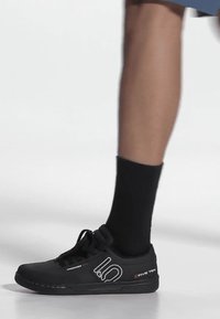 Bein mit schwarzer Socke, das einen schwarzen Five Ten Schnürschuh trägt, auf einer glatten weißen Oberfläche, seitlich betrachtet.