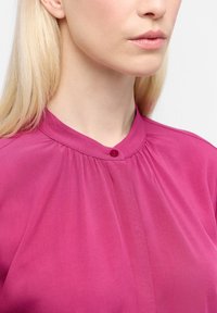 Blusa fucsia con colletto rialzato, caratterizzata da un dettaglio con bottoni e pieghe sottili. Il tessuto è liscio con una leggera lucentezza.