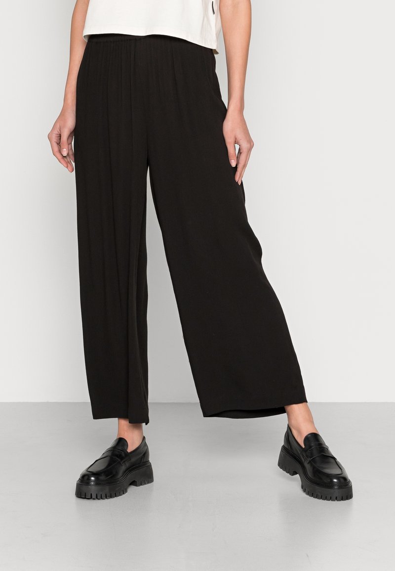Femme portant un pantalon large noir ample et des mocassins noirs épais, debout devant un fond gris clair uni.