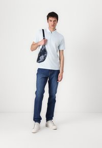 Svijetloplava polo majica, tamnoplave traperice i bijele tenisice. Model drži crno-bijelu torbu s tie-dye uzorkom preko ramena. Jednostavan dizajn, glatka tekstura.