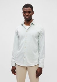 Ανοιχτό πράσινο πουκ άμισο με μακριά μανίκια και κολάρο με κουμπιά, διαθέτει χρυσά κουμπιά και ελαφρώς υφασμάτινη υφή. Φοράει από πάνω ένα λευκό μπλουζάκι.