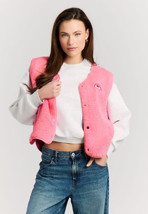 Jonge vrouw die een roze fleecevest draagt over een witte cropped sweatshirt en blauwe spijkerbroek, staand met één hand in de zak tegen een effen achtergrond.
