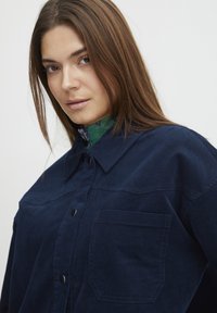 Giacca di velluto a coste blu navy con colletto, taschino sul petto e bottoni a pressione neri. Texture liscia con leggere righe visibili.