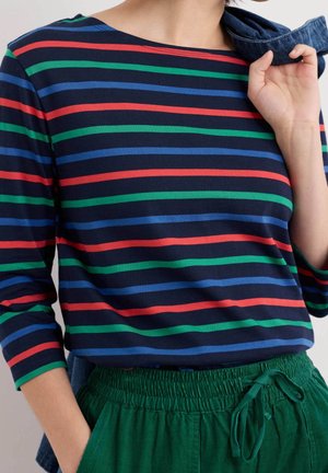 Personne portant une chemise bleu marine avec des rayures horizontales rouges, vertes et bleues et un pantalon vert taille haute avec une taille à cordon noué.