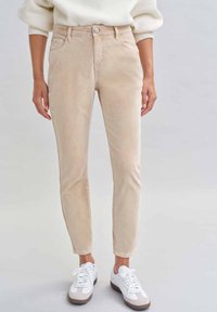Beige Cordhosen mit tailliertem Schnitt, Fronttaschen und Knopfverschluss, kombiniert mit weißen Sneakers und einem strukturierten Pullover.