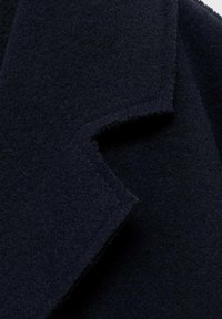 Tejido de abrigo de lana en un azul marino oscuro con una textura suave, que presenta un cuello con muescas y bordes cortados a raw, enfatizando los detalles del diseño minimalista.