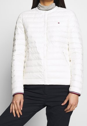 Femme portant une veste matelassée blanche avec des boutons-pression, des poches zippées et un col roulé gris, associée à un pantalon noir.