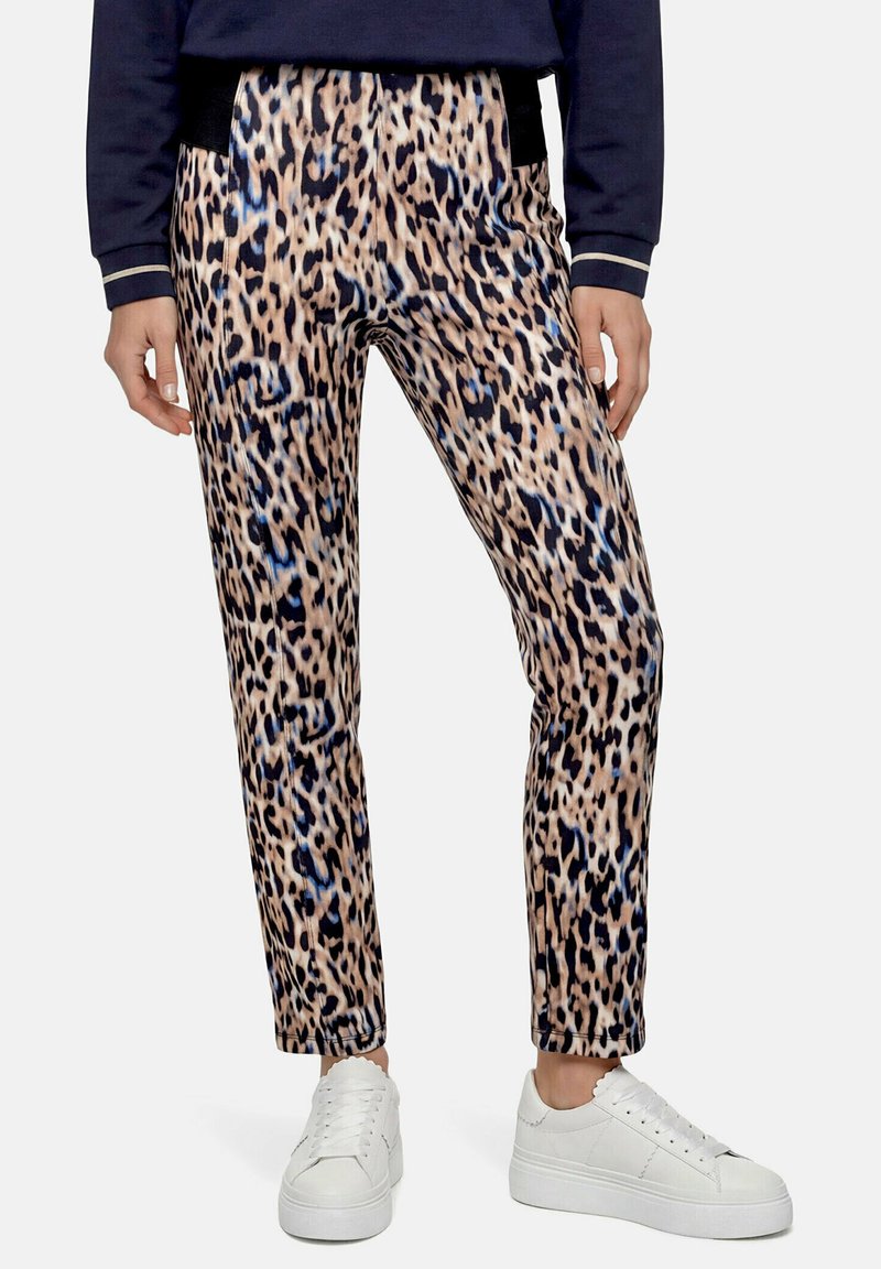 Personne portant un pantalon imprimé léopard, un haut bleu marine et des baskets blanches, debout devant un fond uni.