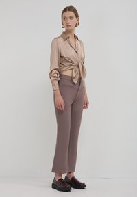 Chemise en satin beige avec un front noué, associée à un pantalon marron ajusté et des mocassins noirs. Texture lisse avec une silhouette cintrée.