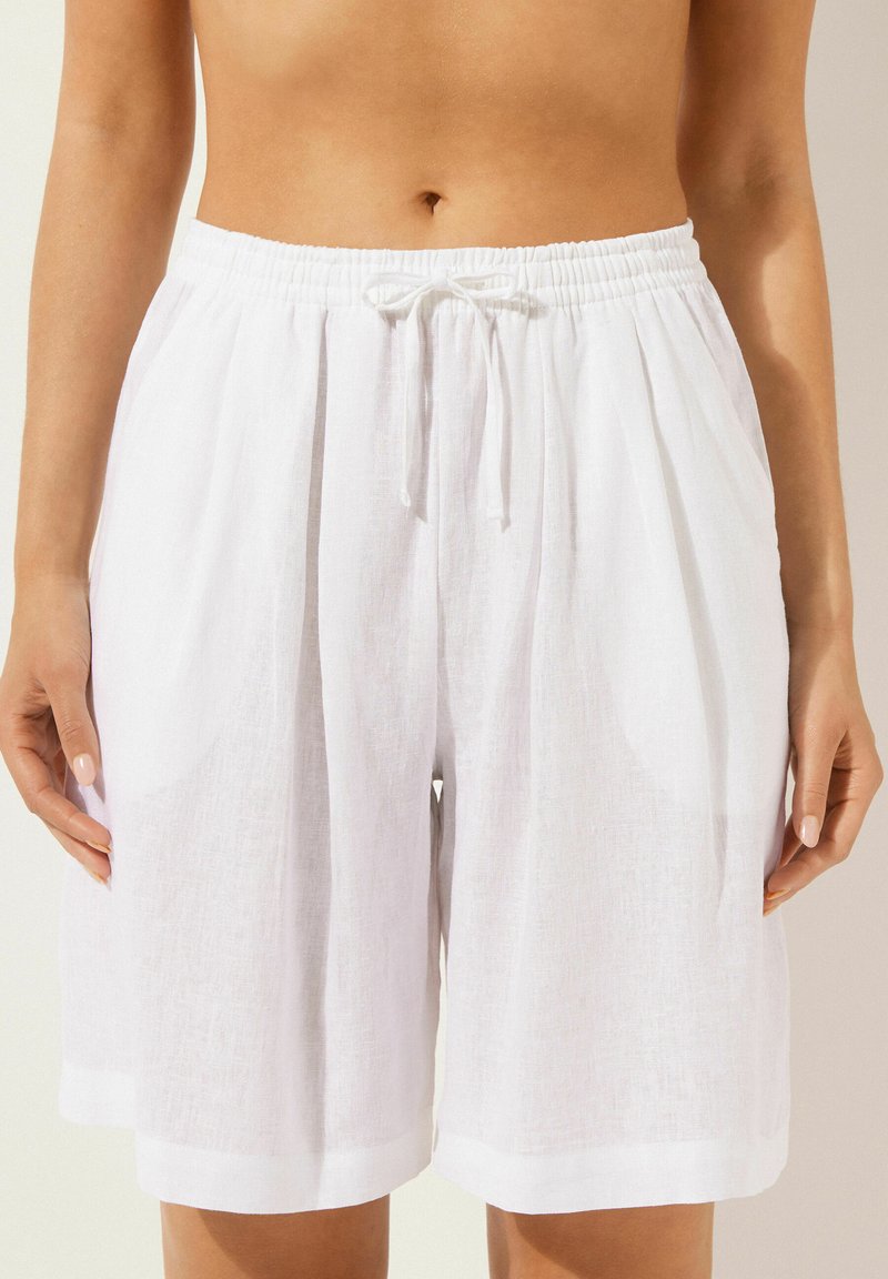 Shorts en lin blancs avec une taille élastique et un cordon de serrage. Présente des poches latérales et une coupe décontractée, avec une texture lisse et sans motifs visibles.