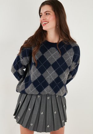 Navy en grijze argyle-gepatroneerde gebreide trui met een geribbelde halslijn, gedragen met een geplooide grijze rok met decoratieve versieringen.