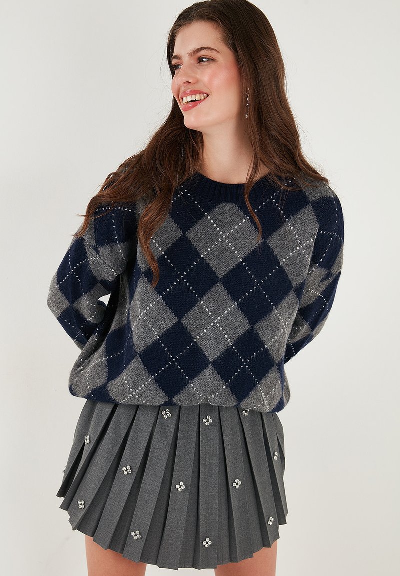 Navy and grey argyle-patterned knit sweater with ribbed neckline paired with a pleated grey skirt featuring decorative embellishments.