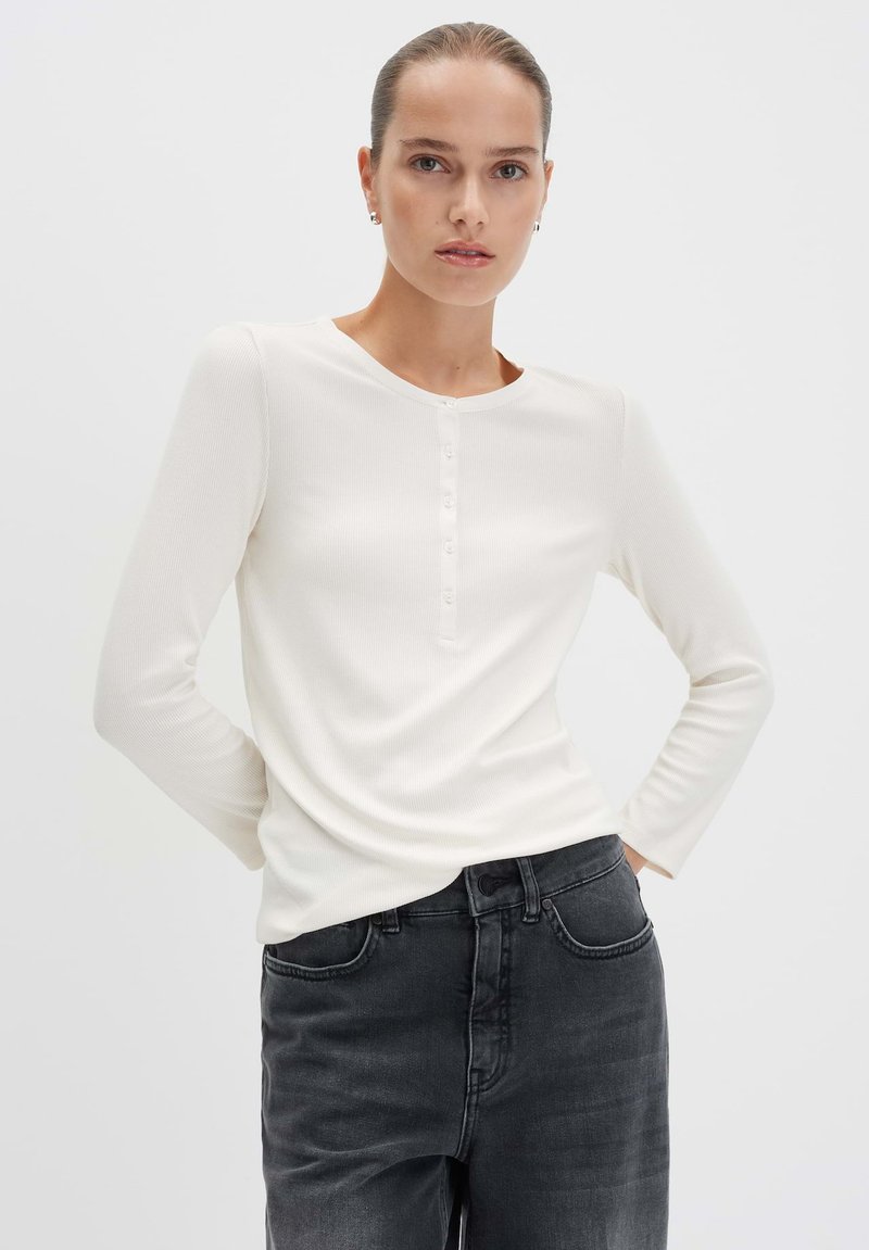 Vrouw met haar in een staart draagt een witte blouse met lange mouwen en donkergrijze high-waisted jeans, staande met haar handen in de zakken.