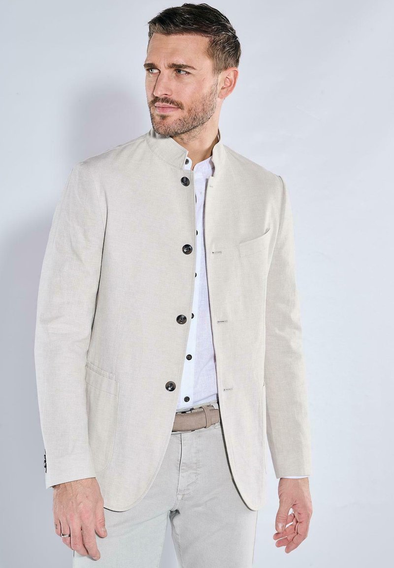 Helle beige Jacke mit Stehkragen, einreihigem Design, schwarzen Knöpfen und seitlichen Taschen. Getragen über einem weißen Hemd und hellen Hosen.