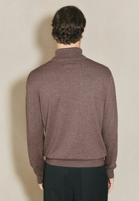 Mann mit lockigem braunem Haar, der einen braunen Rollkragenpullover und schwarze Hosen trägt, steht mit dem Gesicht zu einer beigefarbenen Wand.